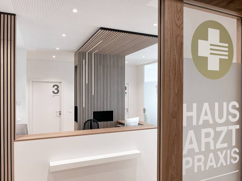 Ein moderner Empfangsbereich einer Hausarztpraxis mit einer Glastür, auf der das Logo und der Schriftzug "Hausarztpraxis" zu sehen sind.