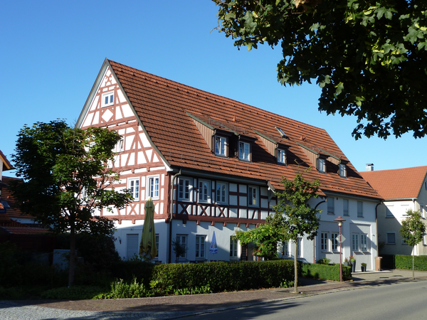 Ein großes Fachwerkhaus mit roten Dachziegeln steht an einer sonnigen Straße, umgeben von Bäumen und Büschen.