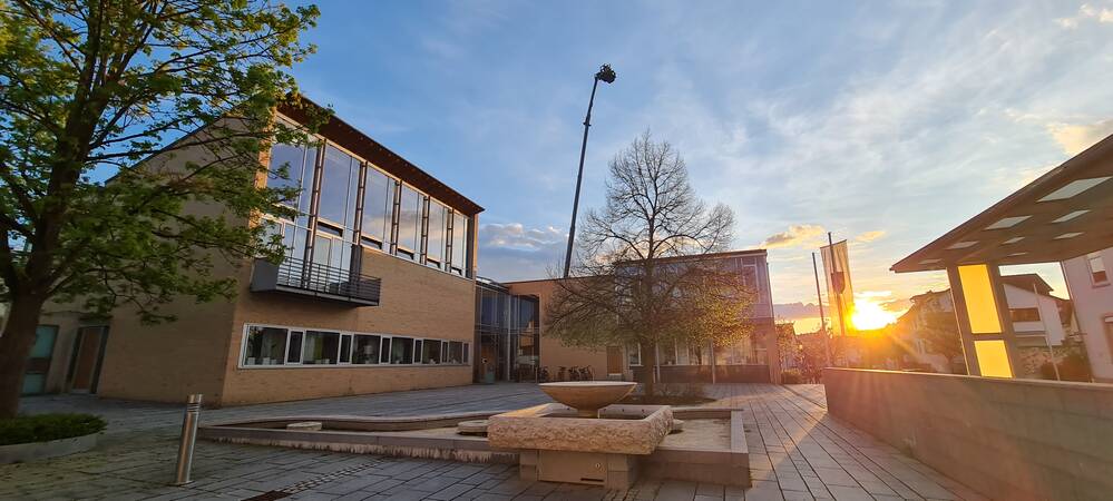 Ein moderner Gebäudekomplex mit großen Fenstern steht im Vordergrund, daneben ein Baum und ein gepflasterter Platz, während die Abendsonne am Horizont untergeht.