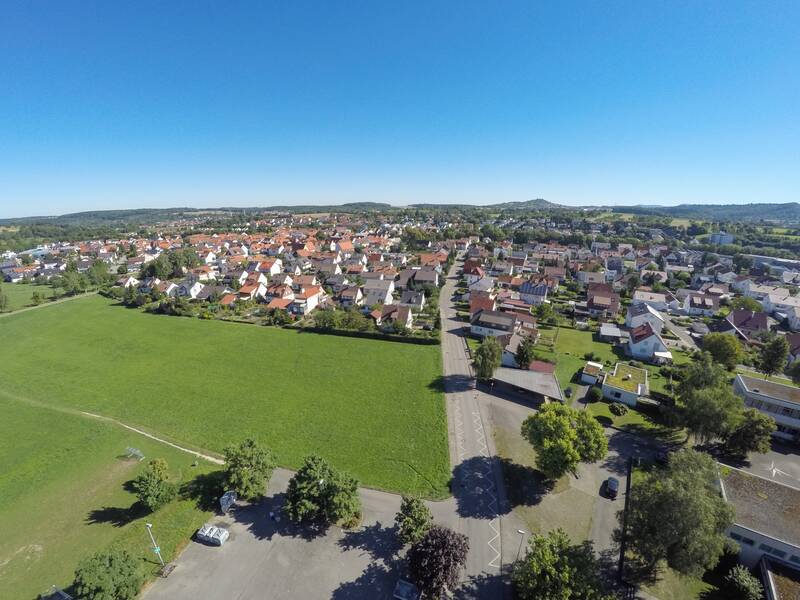 Die Luftaufnahme zeigt eine ländliche Wohngegend mit vielen Häusern, umgeben von grünen Feldern und Bäumen unter einem klaren blauen Himmel.