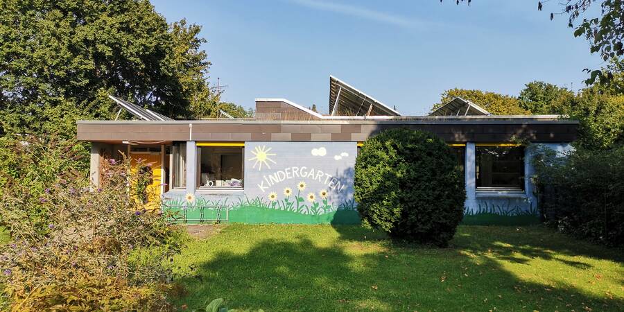 Das Bild zeigt ein einstöckiges Gebäude mit bemalter Fassade und dem Schriftzug "Kindergarten", umgeben von grüner Vegetation und Bäumen, bei sonnigem Wetter.