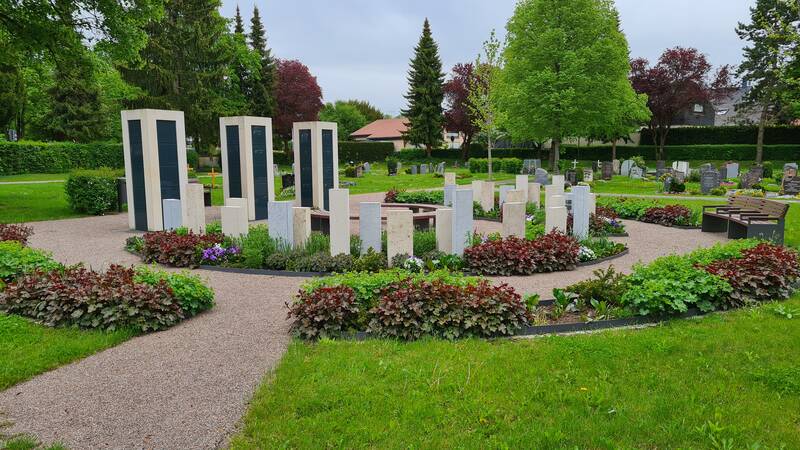Auf dem Bild ist ein gepflegter Friedhof mit einem kreisförmigen Anordnung aus modernen Gedenksteinen, umgeben von Blumenbeeten und Grünflächen zu sehen.