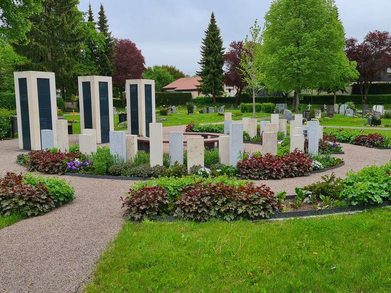Auf dem Bild ist ein gepflegter Friedhof mit einem kreisförmigen Anordnung aus modernen Gedenksteinen, umgeben von Blumenbeeten und Grünflächen zu sehen.