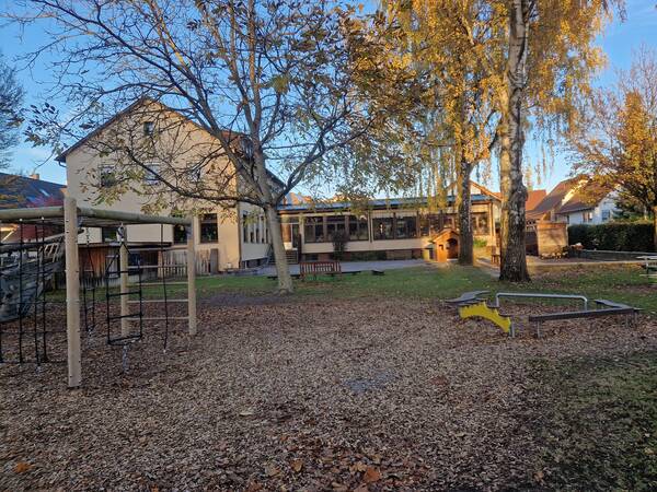 Ein Spielplatz mit verschiedenen Klettergerüsten und Spielgeräten, umgeben von Bäumen und neben einem großen Gebäude, ist in herbstlicher Atmosphäre zu sehen.