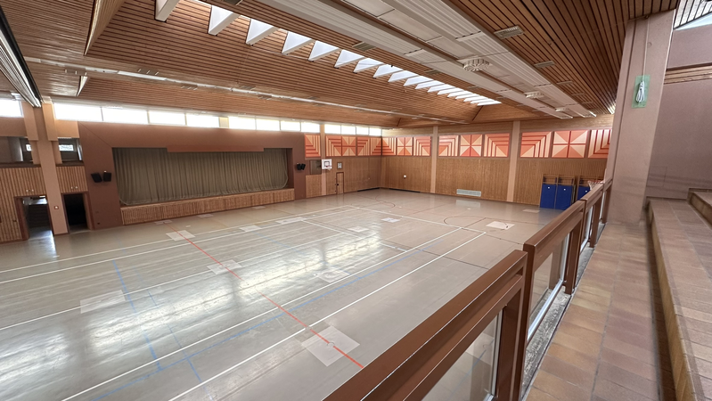Eine geräumige Turnhalle mit Holzwänden und -decke, einem hellen Boden mit Markierungen, und einer Bühne an der Stirnseite.