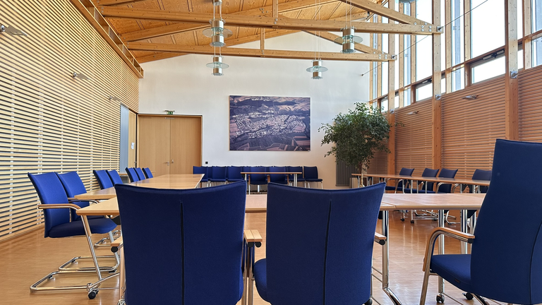 Zu sehen ist ein Besprechungsraum mit einer U-förmigen Anordnung von Tischen mit blauen Stühlen, großen Fenstern und  begrünten Pflanzen.