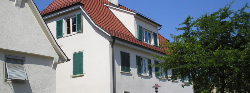 Ein mehrstöckiges, weißes Gebäude mit rotem Dach und kleinen Glockenturm, grünen Fensterläden und einer Treppe am Eingang steht neben einem Baum an einem sonnigen Tag.