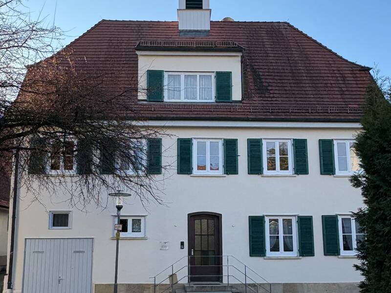 Ein zweistöckiges, weißes Gebäude mit grün umrahmten Fenstern, einem steilen roten Dach und einem kleinen Turm mit Wetterfahne darauf.