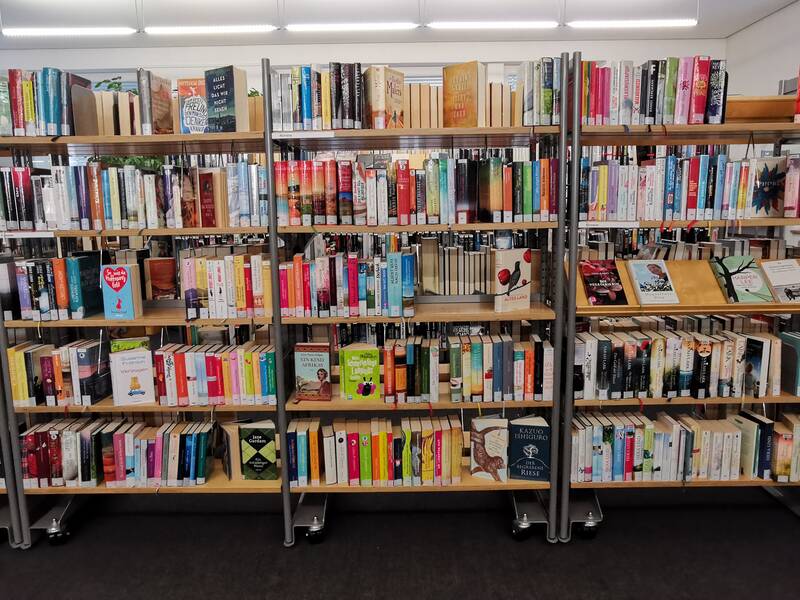 Das Bild zeigt mehrere volle Bücherregale in einer Bibliothek, die mit einer Vielzahl von unterschiedlichen Büchern gefüllt sind.