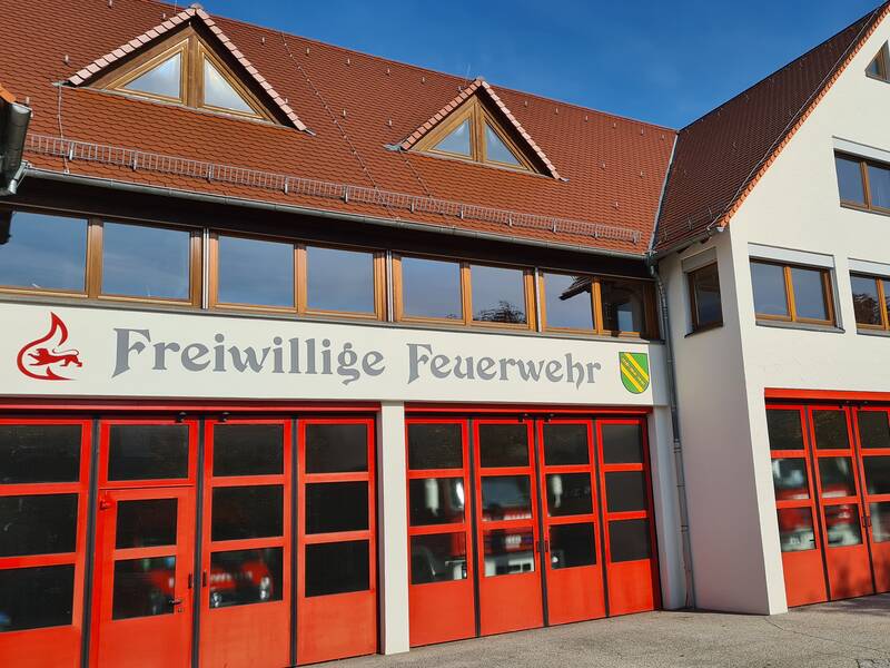 Das Bild zeigt ein Gebäude der Freiwilligen Feuerwehr mit roten Garagentoren und einem Schild, das den Namen der Feuerwehr trägt.