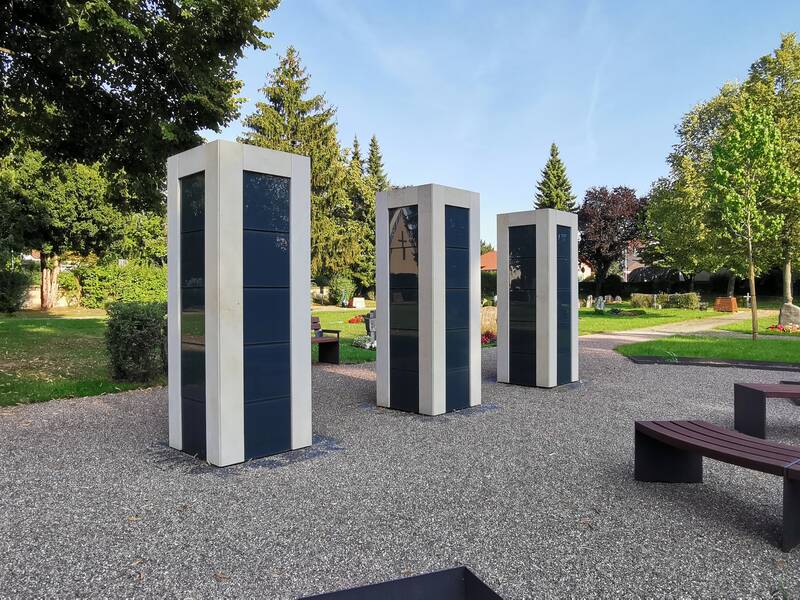 Das Bild zeigt einen Friedhof mit drei modernen Steinkolumbarien aus weißem und dunklem Material, umgeben von Bäumen und Gräbern.