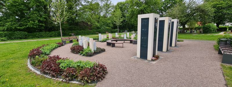 Das Bild zeigt einen kreisförmigen Gedenkort mit mehreren modernen Steinstelen und Bänken, umgeben von grüner Vegetation in einem Park.