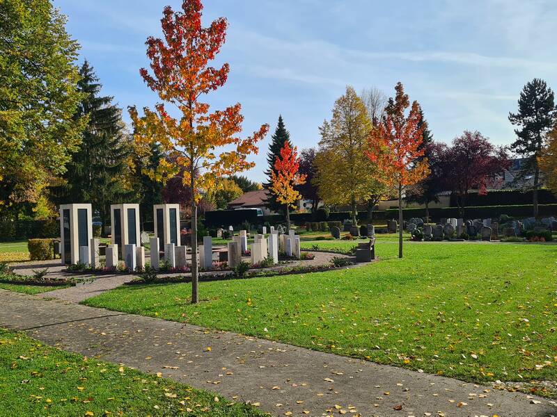 Das Bild zeigt einen kreisförmigen Gedenkort mit mehreren modernen Steinstelen und Bänken, umgeben von grüner Vegetation in einem Park.