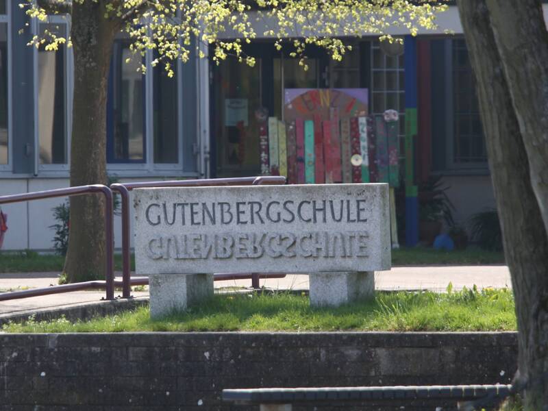 Das Bild zeigt einen Stein mit dem Schriftzug Gutenbergschule Riederich vor dem Pausenhof einer Schule 