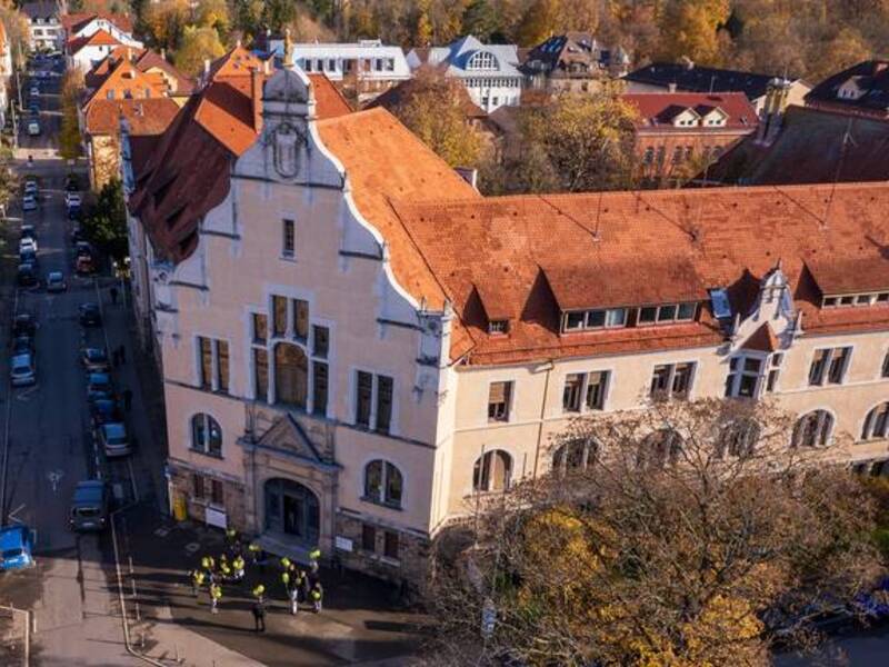 Luftaufnahme eines großen historischen Gebäudes mit rotem Ziegeldach, das in einer herbstlichen, von Bäumen gesäumten Stadtlandschaft liegt.