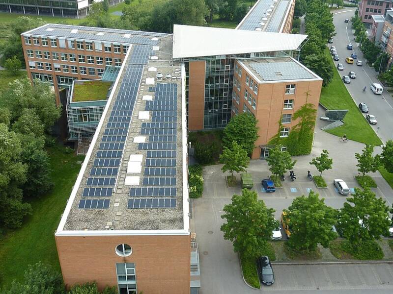 Ein großes, L-förmiges Gebäude mit Solarpanels auf dem Dach, umgeben von Bäumen und geparkten Autos.