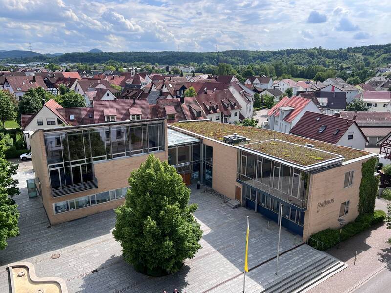 Das Bild zeigt ein modernes Rathausgebäude mit begrüntem Dach und großen Glasfenstern, umgeben von einer kleinen Stadtlandschaft mit roten Ziegeldächern und grünem Hügelland im Hintergrund.