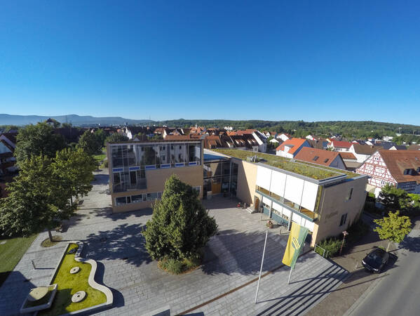 Auf dem Bild ist ein modernes Gebäude mit begrüntem Dach und Glasfassade zu sehen, umgeben von einem gepflasterten Platz mit Bäumen und umgeben von Wohnhäusern.