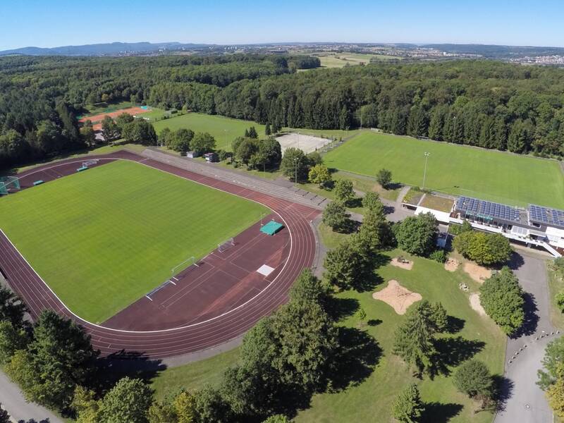 Eine Luftaufnahme zeigt einen weitläufigen Sportkomplex mit einem Fußballplatz, einer Laufbahn und umgeben von Wäldern und Gebäuden.