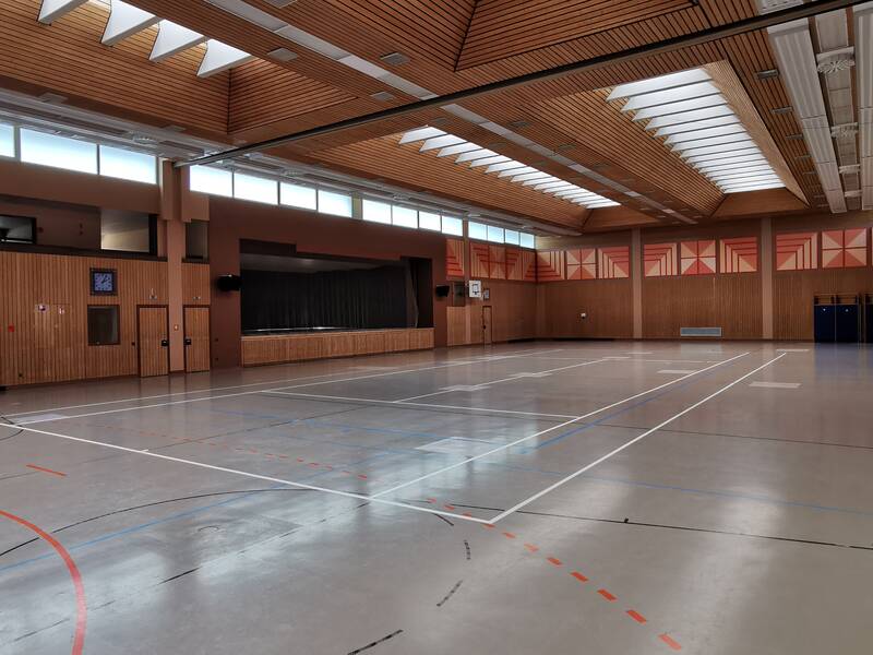 Die Abbildung zeigt eine leere Sporthalle mit Holzwänden, mehreren Basketballkörben und einer großen Fensterfront an der Decke für natürliches Licht.