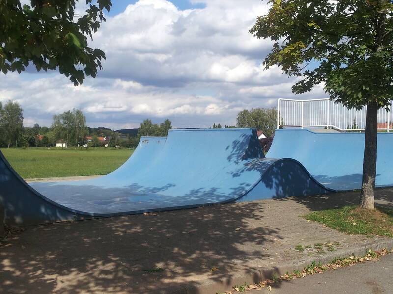 Eine blaue Skateboard-Rampe steht unter Bäumen auf einem Gehweg, mit grüner Wiese und bewölktem Himmel im Hintergrund.