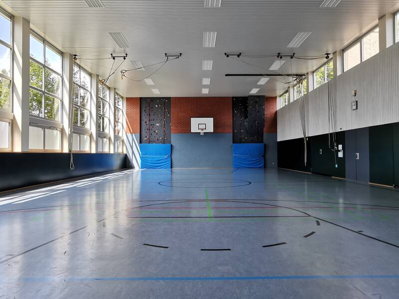 Ein leerer, gut beleuchteter Turnhallenraum mit hohem Dach, großen Fenstern, einer Kletterwand und einem Basketballkorb.