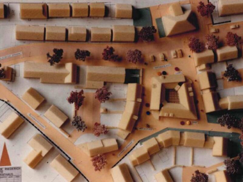 Das Bild zeigt ein architektonisches Modell einer städtischen Planung mit Gebäuden, Straßen und Bäumen.