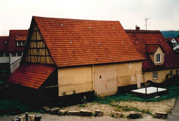 Ein traditionelles Fachwerkhaus mit rotem Ziegeldach steht auf einem Grundstück, umgeben von einem unbefestigten Weg und kleineren Bauten im Hintergrund.