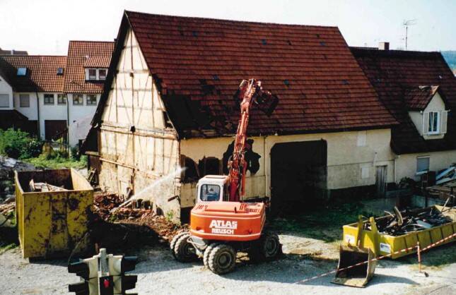 Ein Bagger arbeitet vor einem alten Fachwerkhaus, daneben steht ein großer gelber Container.