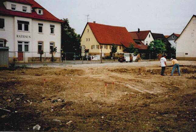 Zwei Personen stehen auf einem unbebauten, erdigen Bauplatz vor einem Gebäude mit der Aufschrift "Rathaus" und weiteren Wohnhäusern im Hintergrund.