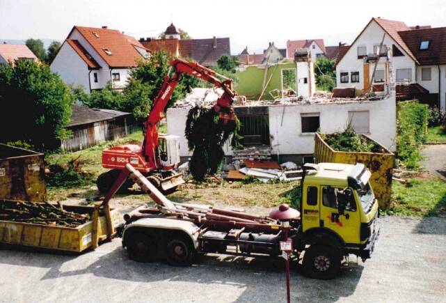 Ein gelber Lastwagen steht vor einem teilweise abgerissenen Gebäude, während ein roter Bagger Schutt hebt, um ihn in einen Container zu laden, umgeben von Wohnhäusern.