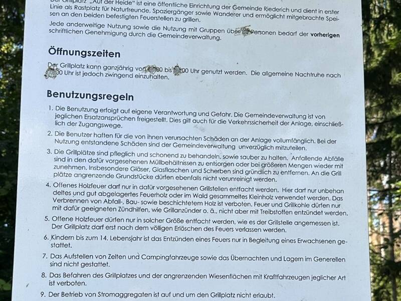 Ein Schild der Gemeinde Riederich informiert über die Regeln und Öffnungszeiten des Grillplatzes „Auf der Heide“ und enthält zusätzlich einige handgeschriebene Kritzeleien.