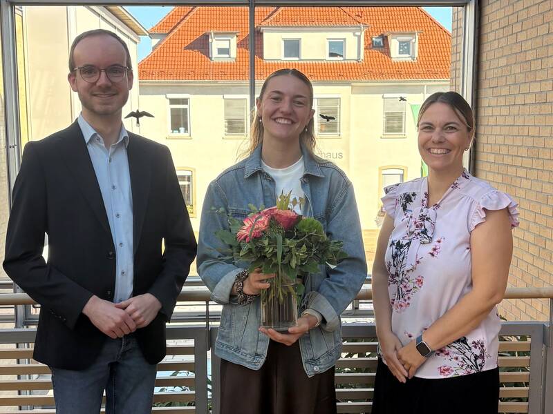 Drei Personen stehen lächelnd auf einem Balkon, wobei die mittlere Person einen Blumenstrauß in den Händen hält. Von links nach rechts Hr. Matthiesen, Frau Gogel, Frau Zunker