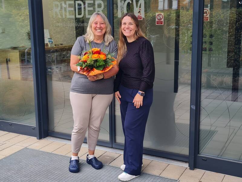 Zwei lachende Frauen stehen vor einem Gebäude, und eine von ihnen hält einen Blumenstrauß.
