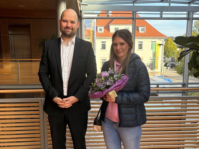 Ein Mann in Anzug steht lächelnd neben einer Frau, die einen Blumenstrauß hält, in einem lichtdurchfluteten Innenraum mit Glasdach.