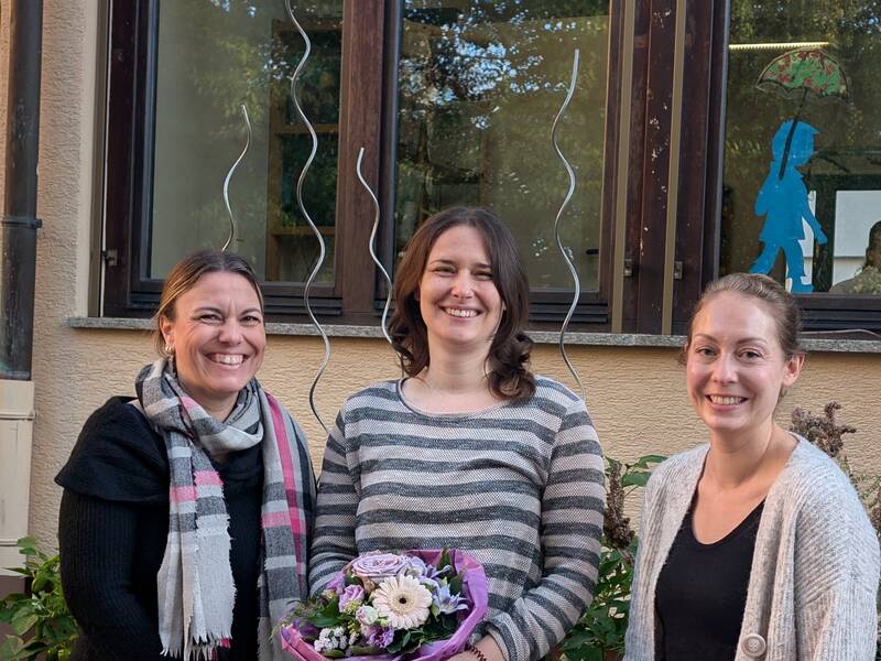 Drei lächelnde Frauen stehen vor einem Gebäude, eine von ihnen hält einen Blumenstrauß.