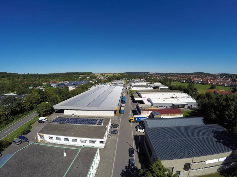 Luftaufnahme eines Industriegebiets mit mehreren großen Hallen und Gebäuden entlang einer Straße, umgeben von grüner Landschaft und einem entfernt gelegenen Wohngebiet.