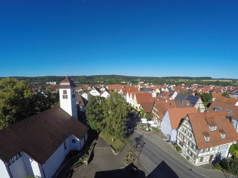 Die Luftaufnahme zeigt ein malerisches Dorf mit einer Kirche, Fachwerkhäusern und Bäumen unter einem klaren blauen Himmel.